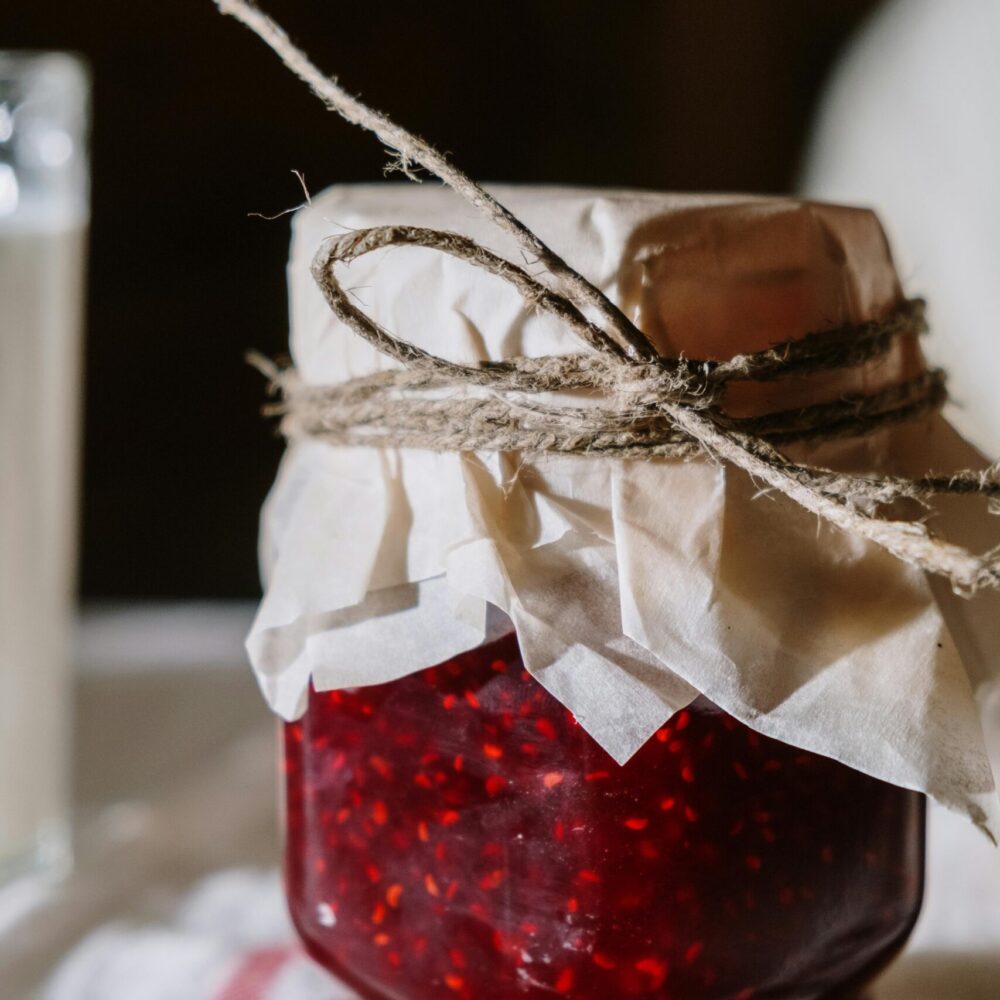 A close-up of homemade raspberry jam in a rustic glass jar on a white table.